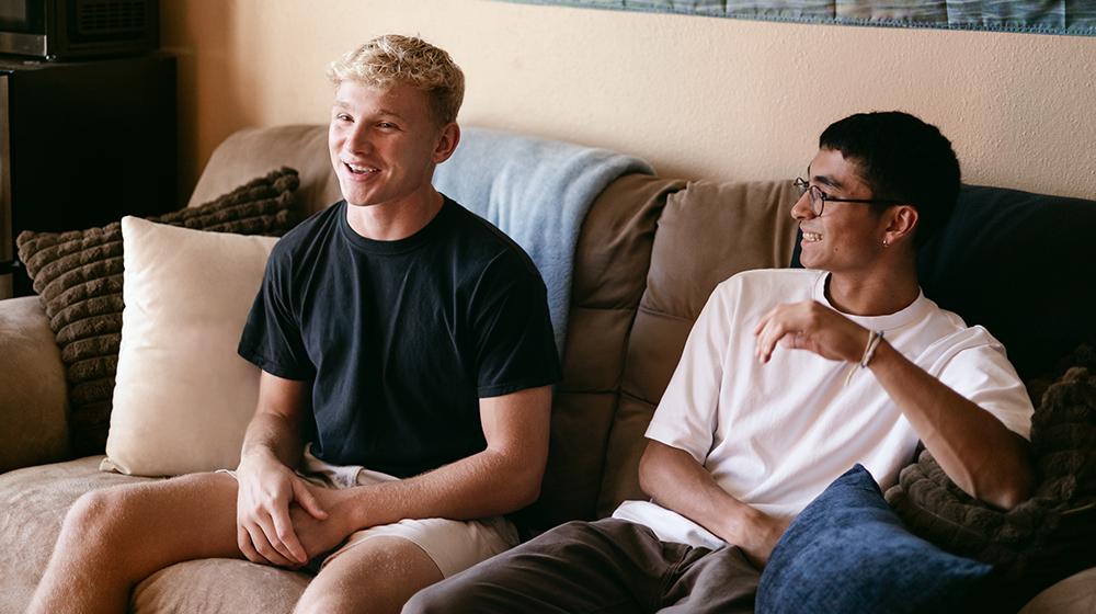 Two young men sitting on a couch, smiling and talking in a relaxed setting.